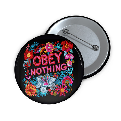 A black round pinback button featuring floral embroidery-style designs and bold red text that reads "Obey Nothing."